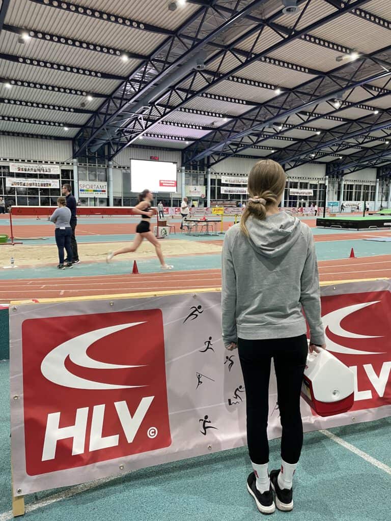 weibliche Person von hinten, die in einer Leichtathletikhalle an der Laufband steht. In ihrer Hand hält sie eine Kryobox.